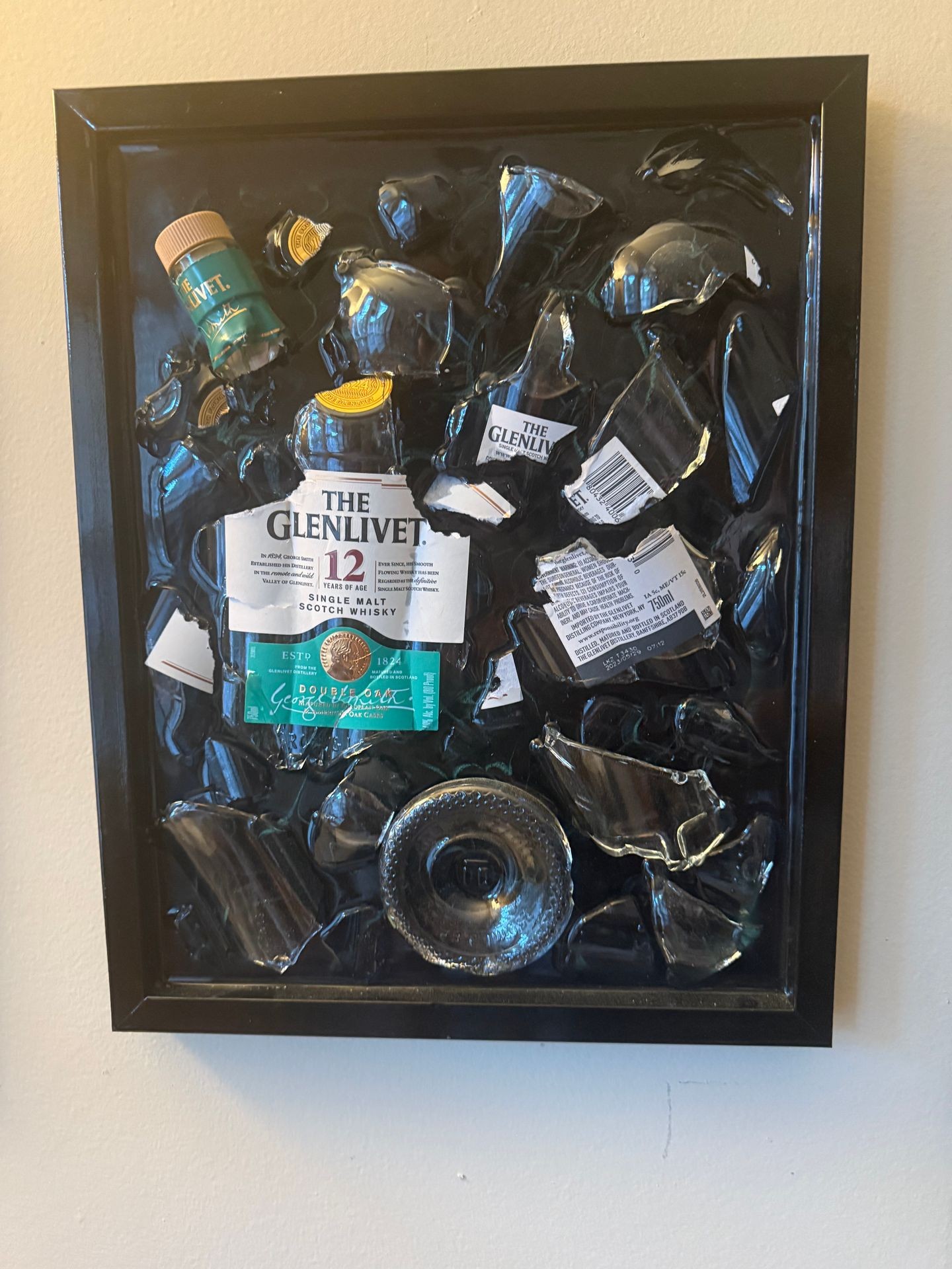 Framed display of shattered Glenlivet bottle pieces and labels on a wall.